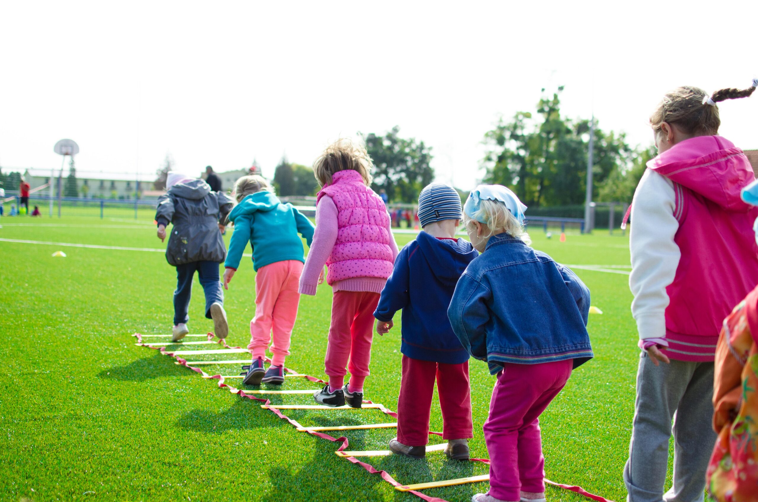 Children enjoy an outdoor activity on a grassy field, stepping over a ladder.