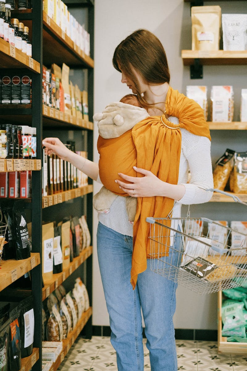 A mother wearing a baby in a sling selects products in a grocery store, holding a basket.